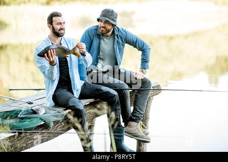 Zwei glückliche Fischer holding Fisch sitzen auf dem Holzsteg beim Fischen auf dem See am Morgen Stockfoto