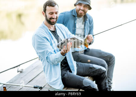 Zwei glückliche Fischer holding Fisch sitzen auf dem Holzsteg beim Fischen auf dem See am Morgen Stockfoto