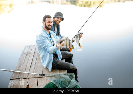 Zwei glückliche Fischer holding Fisch sitzen auf dem Holzsteg beim Fischen auf dem See am Morgen Stockfoto