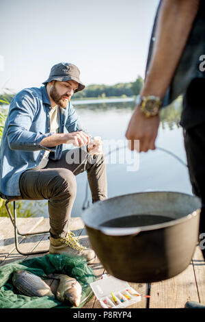 Zwei Fischer bereitet etwas zu Essen während der Picknick auf der hölzernen Pier in der Nähe des Sees am Morgen Stockfoto