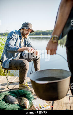 Zwei Fischer bereitet etwas zu Essen während der Picknick auf der hölzernen Pier in der Nähe des Sees am Morgen Stockfoto