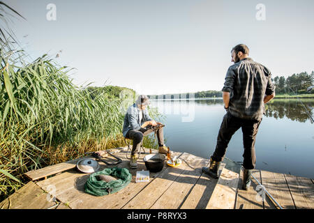 Zwei Fischer entspannen während das Picknick auf der hölzernen Pier in der Nähe des Sees am Morgen Stockfoto