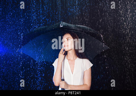 Junge Frau unter dem Dach. Schutz vor Regen. Brunette Mädchen Holding ein Regenschirm. Portrait im Studio auf schwarzem Hintergrund. Konzept der Optimismus Stockfoto