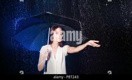 Junge Frau unter dem Dach. Schutz vor Regen. Brunette Mädchen Holding ein Regenschirm. Portrait im Studio auf schwarzem Hintergrund. Konzept der Optimismus Stockfoto