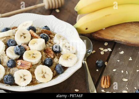 Warmes Frühstück der gesunden Hafermehl mit Pekannüssen, Bananen, Blaubeeren und Honig über eine rustikale Hintergrund. Bild geschossen von Overhead. Stockfoto