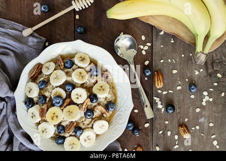Warmes Frühstück der gesunden Hafermehl mit Pekannüssen, Bananen, Blaubeeren und Honig über eine rustikale Hintergrund. Bild geschossen von Overhead. Stockfoto