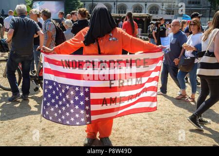 London, Großbritannien. 13. Juli 2018. Zehntausende von Menschen auf den Straßen von London gegen uns Präsident Donald Trump Besuch in Großbritannien zu protestieren. Im Bild: Eine mit Kapuze und orange Overall Figur hält eine invertierte US-Flagge mit den Worten Unabhängigkeit von Amerika über Es Stenciled. Credit: Mark Phillips/Alamy leben Nachrichten Stockfoto
