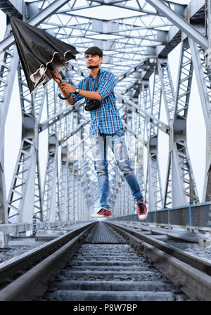Junge Erwachsene indischer Junge schwebend auf railroad Track mit Regenschirm in der hand und die kamera auf den Hals unter Metal Bridge Struktur. Stockfoto