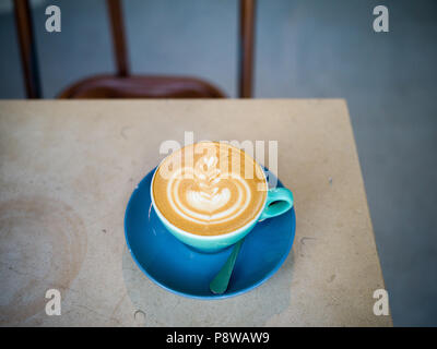 Tasse Kaffee auf Tisch mit schönen latte Kunst, flache weiße, blaue Schale mit einer halben Stuhl im Hintergrund. Keine Personen oder Marke in Bild. Sauber und einfach Stockfoto