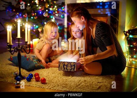 Junge Mutter und ihren zwei kleinen Töchtern eine zauberhafte Weihnachten Geschenk von einem Weihnachtsbaum im gemütlichen Wohnzimmer im Winter Stockfoto