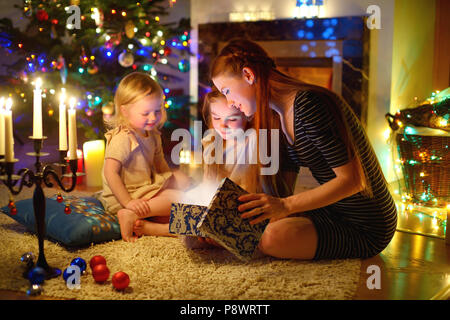 Junge Mutter und ihren zwei kleinen Töchtern eine zauberhafte Weihnachten Geschenk von einem Weihnachtsbaum im gemütlichen Wohnzimmer im Winter Stockfoto