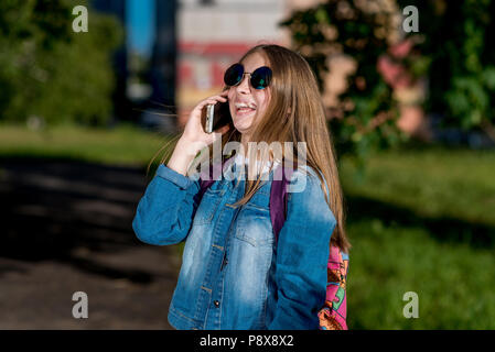 Mädchen Schülerin sprechen Smartphone. Im Sommer, in Jeans Bekleidung, Rucksack, Sonnenbrille ist hinter ihr zurück. Er lacht Hell und emotional. Lächelt und genießt das Gespräch. Stockfoto