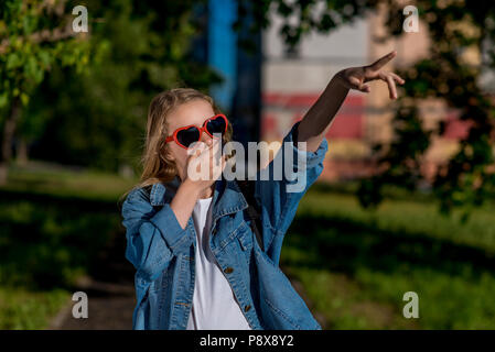 Mädchen Schulmädchen. Sommer in der Natur. Sonnenbrillen in der Form des Herzens. Die Finger seiner Hand Punkte zu Seite. Das Konzept der überraschen lassen Sie sich überraschen. Gefühl der Freude Empörung. Stockfoto