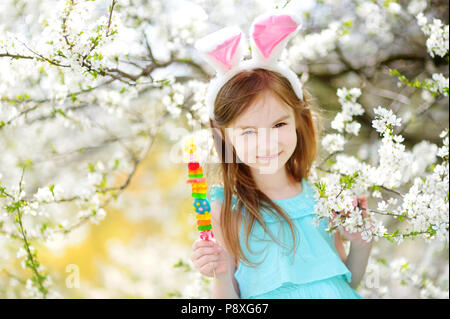 Adorable kleine Mädchen mit Hasenohren Essen bunte Bonbons Kaugummi auf einem Stock in Blooming Cherry Garden am Ostersonntag Stockfoto