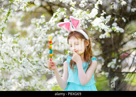 Adorable kleine Mädchen mit Hasenohren Essen bunte Bonbons Kaugummi auf einem Stock in Blooming Cherry Garden am Ostersonntag Stockfoto