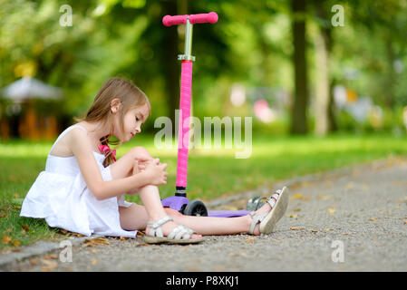 Kleines Mädchen auf dem Boden sitzend, nachdem Sie fiel, während das reiten Ihr scooter am Sommer, Park Stockfoto