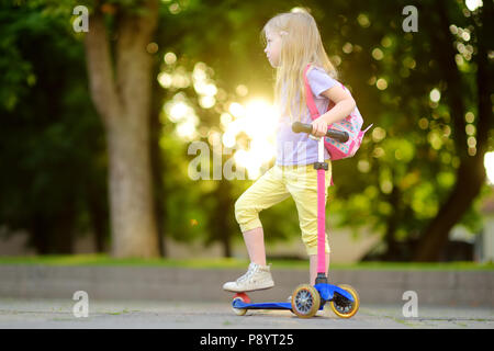 Kleines Kind lernen einen Roller in einem Stadtpark an sonnigen Sommerabend zu fahren. Süße kleine Mädchen reiten eine Rolle. Aktive Freizeit und Outdoor Sport für Stockfoto