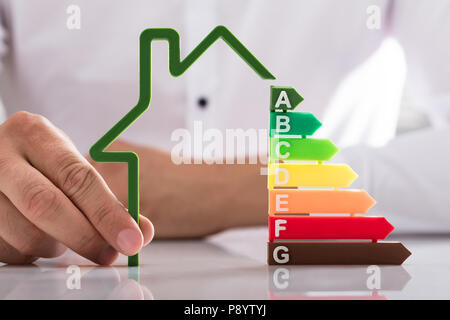 Der Geschäftsmann, der Hand, die Umrisse des Hauses Modell mit Energieeffizienz Rate auf reflektierenden Schreibtisch Stockfoto