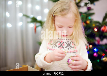 Adorable kleine Mädchen schmücken einen Weihnachtsbaum mit bunten Christbaumkugeln aus Glas zu Hause Stockfoto