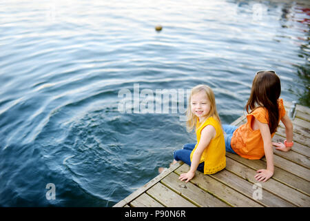 Zwei süße kleine Mädchen sitzen auf einer hölzernen Plattform durch den Fluss oder See Ihre Füße eintauchen in das Wasser an warmen Sommertagen Stockfoto