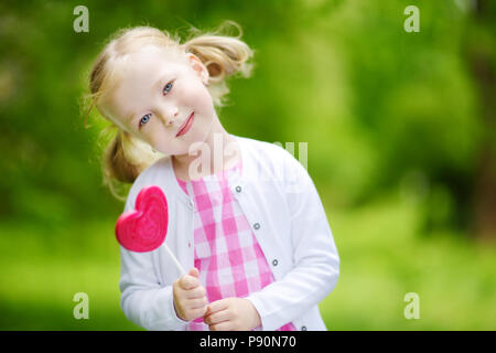 Süße kleine Mädchen mit großen herzförmigen Lollipop im Freien auf schönen Sommertag. Kind mit Lolly candy draußen. Stockfoto