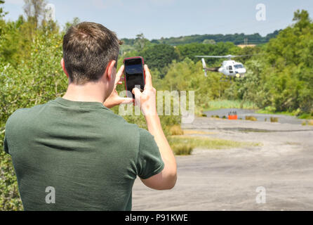 Pontypridd, Wales, 14. Juli 2018. Pontypridd, Wales, 14. Juli 2018. Person, die ein Bild mit einem Handy von einem Hubschrauber verwendet werden, um ein Feuer zu bekämpfen. Der Helikopter ist mit der Maus über einem Teich, seinen Eimer mit Wasser zu füllen. Der Hubschrauber wird verwendet, um ein Gras, das Feuer auf die graig Berghang oberhalb Pontypridd in South Wales zu kämpfen. Es zieht Wasser aus einem Teich auf dem Gelände der ehemaligen Cwm Koksöfen und Zeche Beddau in der Nähe von Pontypridd. Bild 14. Juli 2018. Quelle: Ceri Breeze/Alamy Leben Nachrichten genommen Stockfoto