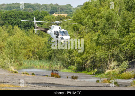 Pontypridd, Wales, 14. Juli 2018. Kampf gegen Gras Brände mit einem Hubschrauber. Staub auf dem Boden ist, peitschte wie ein Hubschrauber aus Aufzüge mit einem Eimer Wasser. Der Hubschrauber wird verwendet, um ein Gras, das Feuer auf die graig Berghang oberhalb Pontypridd in South Wales zu kämpfen. Der Helikopter ist die Abholung von Wasser aus einem Teich auf dem Gelände der ehemaligen Cwm Koksöfen und Zeche Beddau in der Nähe von Pontypridd. Bild 14. Juli 2018. Quelle: Ceri Breeze/Alamy Leben Nachrichten genommen Stockfoto