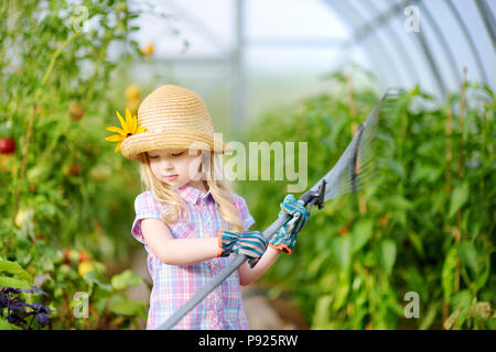 Adorable kleine Mädchen mit Strohhut und Kinder Garten Handschuhe mit ihr spielen Spielzeug Garten Werkzeuge in einem Gewächshaus auf sonnigen Sommertag Stockfoto