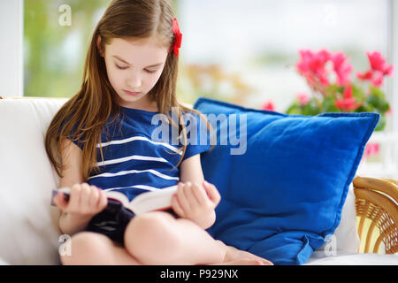 Adorable kleine Mädchen das Lesen eines Buches in Weiß Wohnzimmer auf schönen Sommertag. Smart Schülerin ihre Hausaufgaben zu Hause nach der Schule. Stockfoto