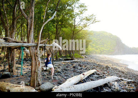 Junge weibliche Touristen entspannen auf einer handgemachten Schwingen auf felsigen Strand von Pololu Tal auf der grossen Insel von Hawaii, USA Stockfoto
