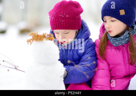 Zwei Entzückende kleine Mädchen einen Schneemann bauen gemeinsam im schönen Winter Park. Süße Schwestern Spielen im Schnee. Winter Aktivitäten für Kinder. Stockfoto