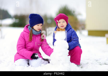 Zwei Entzückende kleine Mädchen einen Schneemann bauen gemeinsam im schönen Winter Park. Süße Schwestern Spielen im Schnee. Winter Aktivitäten für Kinder. Stockfoto