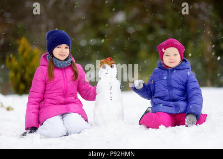 Zwei Entzückende kleine Mädchen einen Schneemann bauen gemeinsam im schönen Winter Park. Süße Schwestern Spielen im Schnee. Winter Aktivitäten für Kinder. Stockfoto