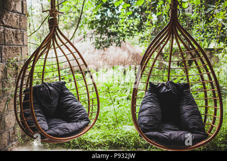 Zwei rattan hängenden Korbstühlen mit schwarzem Kissen in Garten, Sommer Stockfoto