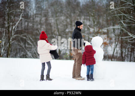 Zwei entzückenden kleinen Mädchen und ihrem Vater ein Schneemann zusammen Gebäude in schöner Winter Park. Süße Schwestern Spielen im Schnee. Aktivitäten im Winter für Stockfoto