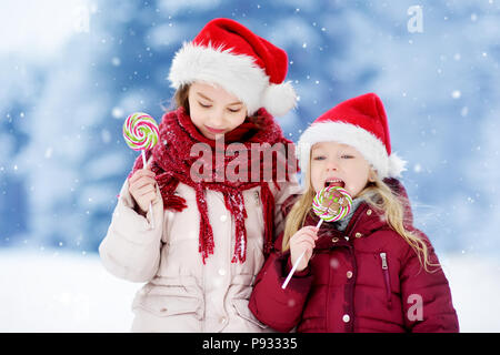 Zwei Entzückende kleine Schwestern tragen Nikolausmützen in riesigen gestreiften Weihnachten Lutscher auf schönen Wintertag. Kinder mit Lolly Bonbons auf Weihnachten da Stockfoto