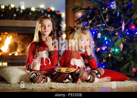 Glückliche kleine Schwestern in Milch und Cookies durch einen Kamin in der gemütlichen dunklen Wohnzimmer am Heiligabend. Feiern Weihnachten zu Hause. Stockfoto