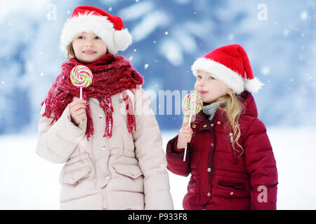 Zwei Entzückende kleine Schwestern tragen Nikolausmützen in riesigen gestreiften Weihnachten Lutscher auf schönen Wintertag. Kinder mit Lolly Bonbons auf Weihnachten da Stockfoto