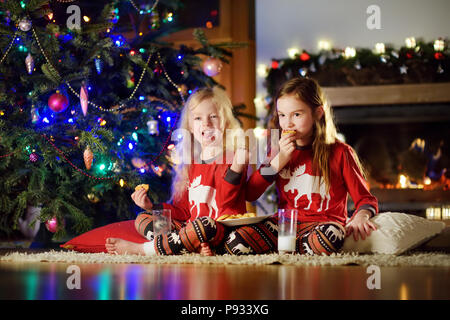 Glückliche kleine Schwestern in Milch und Cookies durch einen Kamin in der gemütlichen dunklen Wohnzimmer am Heiligabend. Feiern Weihnachten zu Hause. Stockfoto
