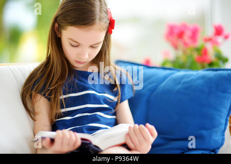 Adorable kleine Mädchen das Lesen eines Buches in Weiß Wohnzimmer auf schönen Sommertag. Smart Schülerin ihre Hausaufgaben zu Hause nach der Schule. Stockfoto