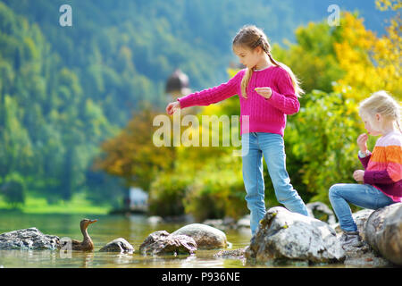 Adorable Schwestern spielen durch Konigssee See in Deutschland an einem warmen Sommertag. Süße Kinder Spaß Fütterung Enten und warfen Steine in den See. Su Stockfoto