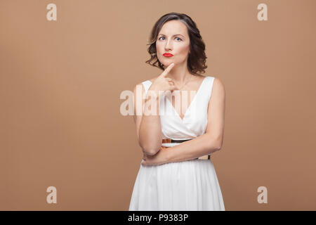 Schöne Frau mittleren Alters denken und verwirrt. Emotionale Ausdruck Frau im weißen Kleid, roten Lippen Make-up und dunkle lockige Frisur. Studio shot, in Stockfoto