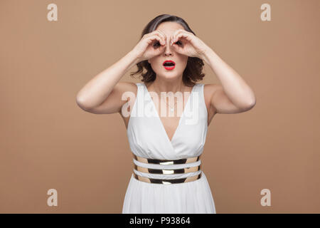 Frau mittleren Alters weit weg auf der Suche nach Fernglas Geste. Emotionale Ausdruck Frau im weißen Kleid, roten Lippen und dunkle lockige Frisur. Studio shot, ich Stockfoto