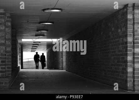 Zwei Personen Silhouette gegen ein helles Licht zu Fuß durch einen Tunnel mit gemauerten Wänden auf die Kamera (Schwarzweiß) Stockfoto