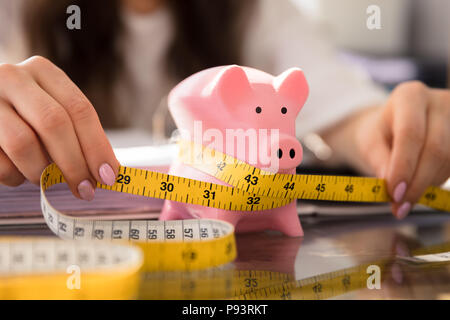 Die Geschäftsfrau Hand messen Sparschwein mit gelbem Maßband auf Glas Schreibtisch Stockfoto