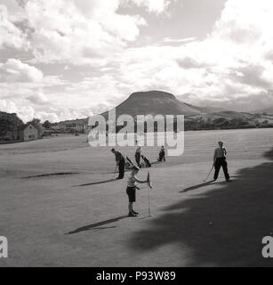 1950, historische, Golf in Co Down, Nordirland, ein Junge Caddy hält die Flagge auf dem Grün als Golfspieler bereiten zu putten. Die schöne Mourne Mountains im Hintergrund. Stockfoto