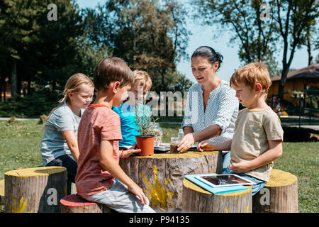 Gruppe von Schulkindern mit einer Lektion in einem Park. Kinder sitzen um einen Tisch und Lernens zusammen mit einem Lehrer im Garten. Stockfoto