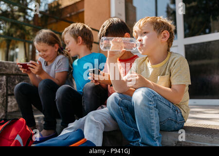 Junge Schüler sitzen auf der Treppe außerhalb der Schule spielen mit Handys während der Schulferien. Primäre Studierende vor der Schule ausruhen. Stockfoto