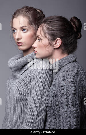 Zwei junge Frauen, die in grauen pullover auf Grau studio Hintergrund. Schöne Mädchen stretching Hände in der Umarmung. Weibliche Freundschaft Konzept. Stockfoto