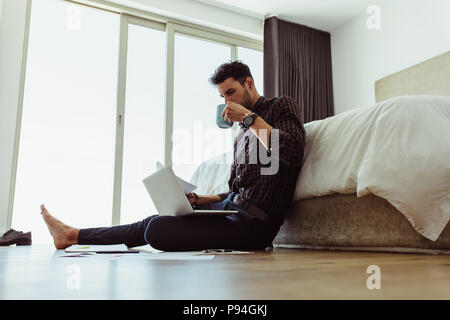 Freelancer Papier in der Hand halten, während auf Laptop zu Hause arbeiten. Mann bei der Arbeit am Laptop sitzen auf dem Boden trinken Kaffee. Stockfoto